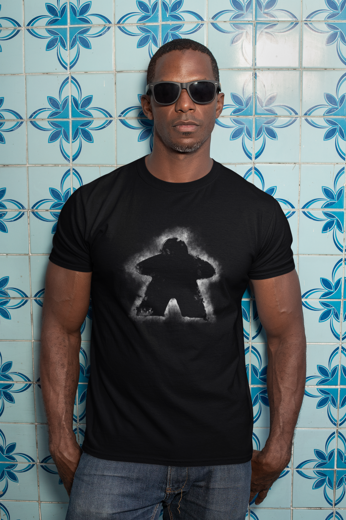 Man wearing a black t-shirt with a glowing meeple design against a blue tiled wall.
