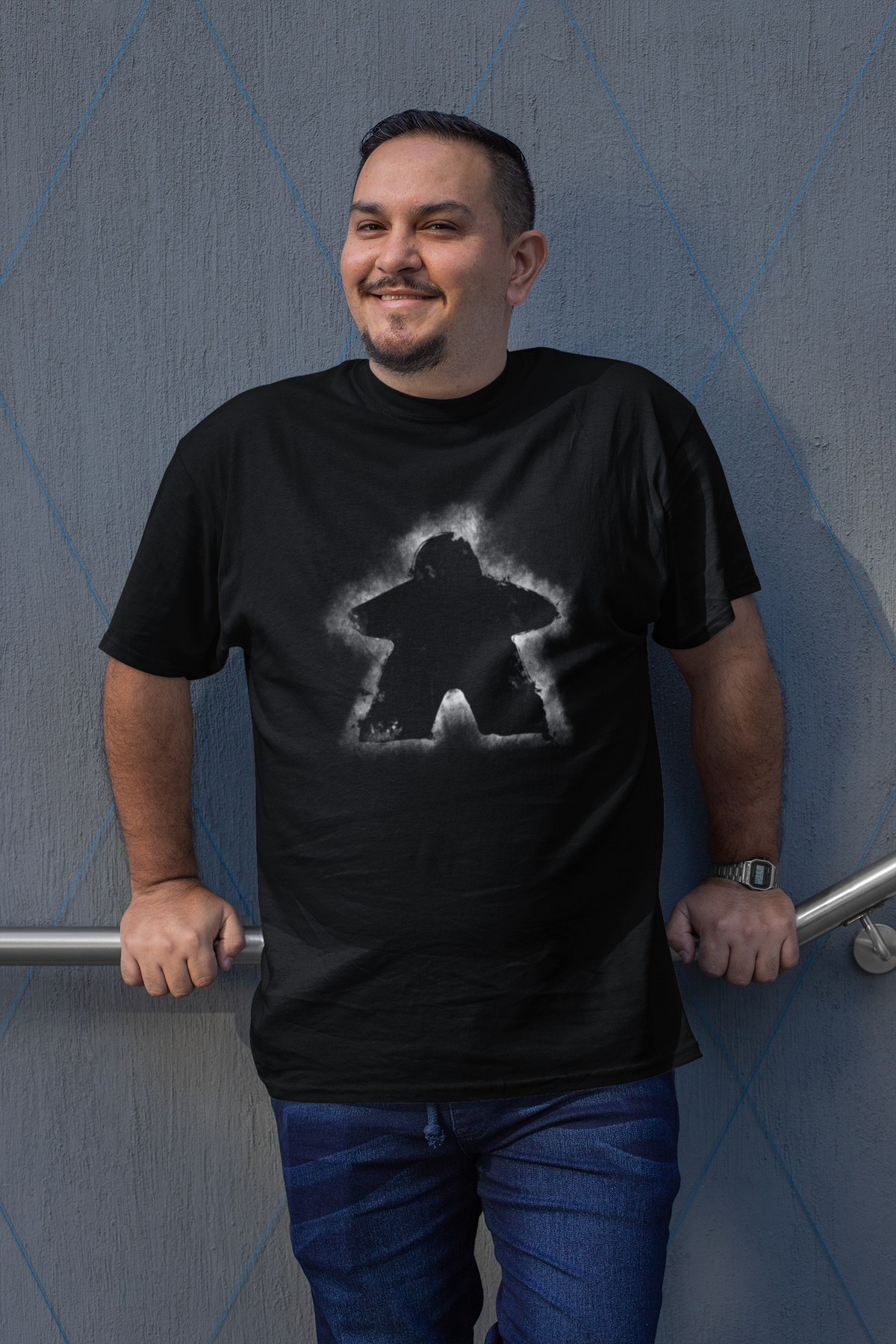 Man wearing a black t-shirt with a glowing meeple design against a blue wall.