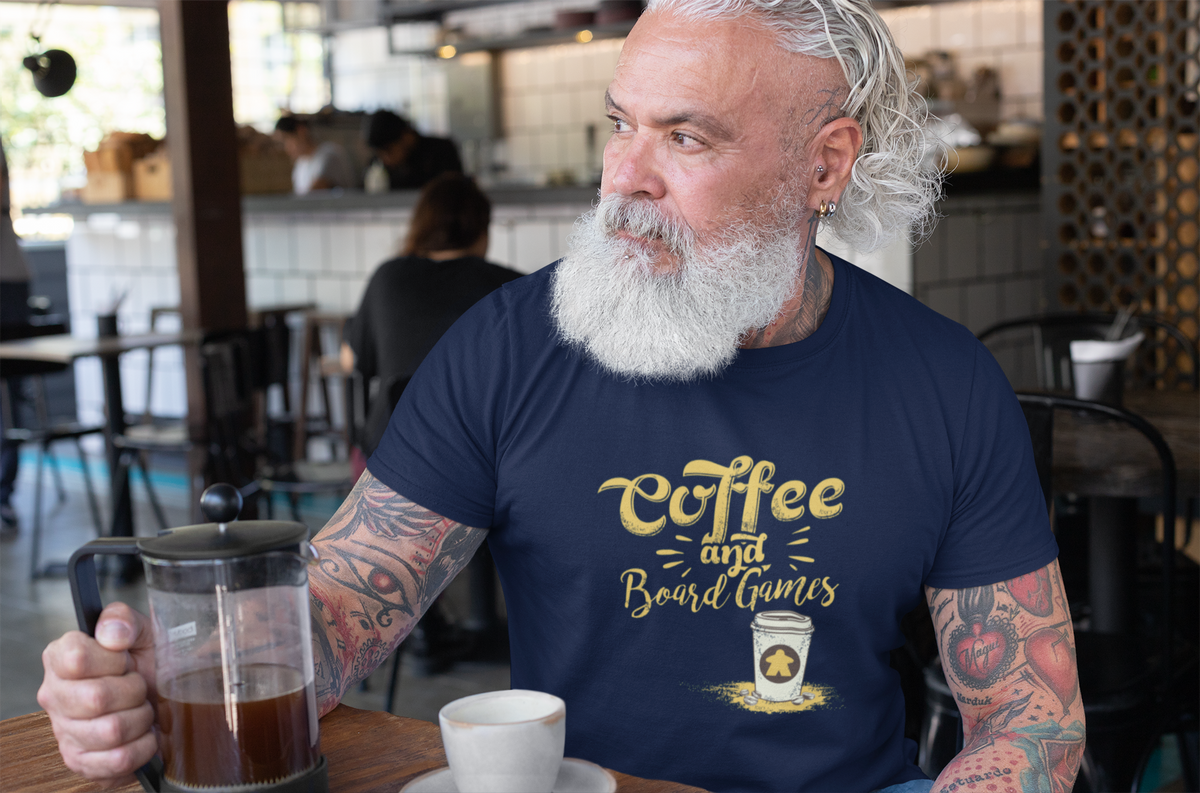 Older Man with tattoos wearing a 'Coffee and Board Games' t-shirt in a cafe.