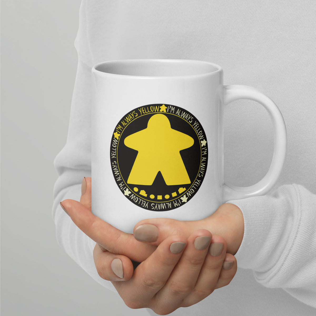 A hand holding a white ceramic mug with our I'm Always Yellow meeple design 