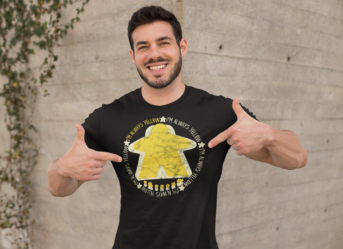 Man wearing a black t-shirt with our I'm Always Yellow Meeple graphic design, pointing at the shirt.