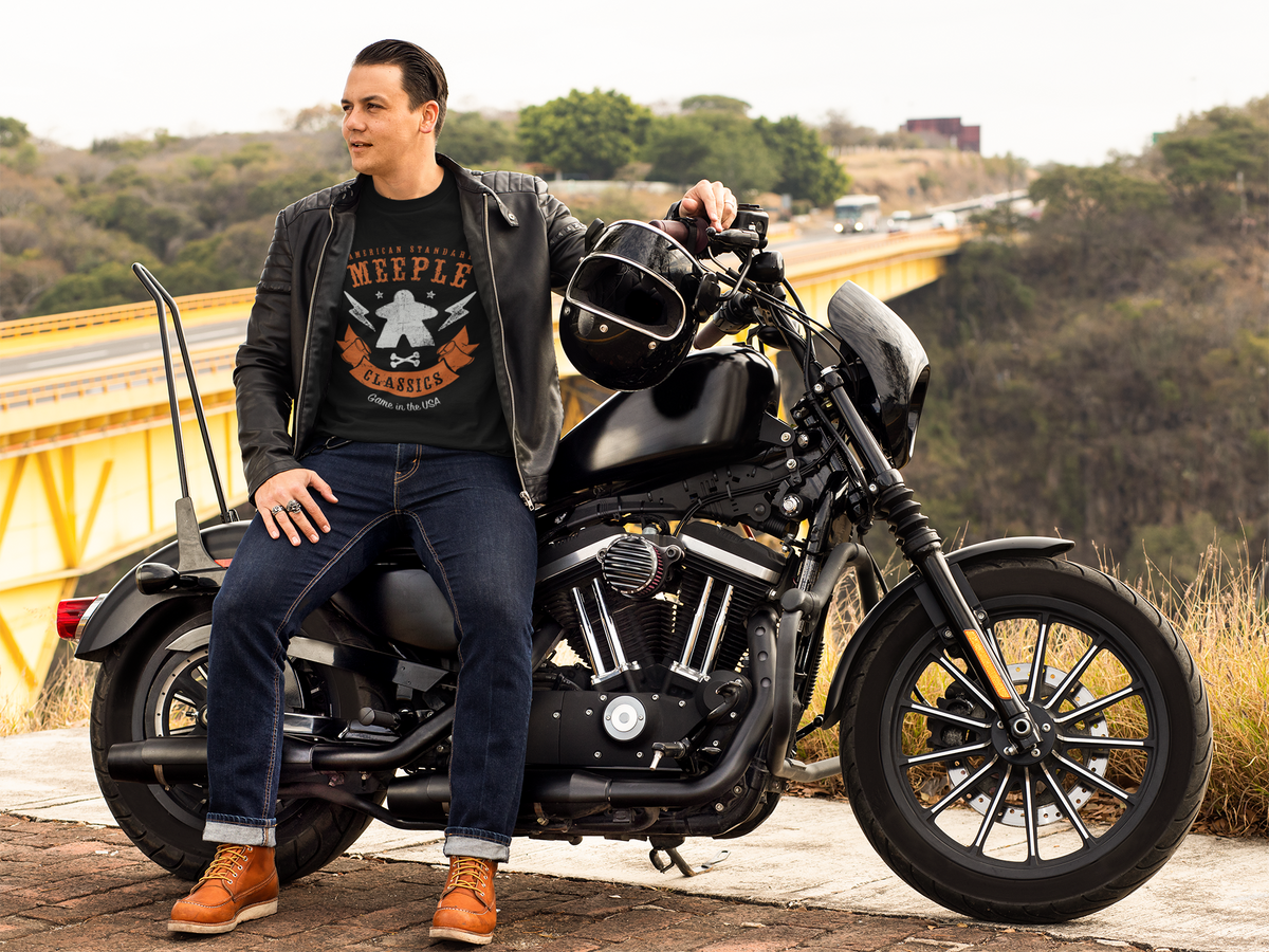 Man sitting on a motorcycle with a scenic background wearing American Standard Meeple Classics T-Shirt
