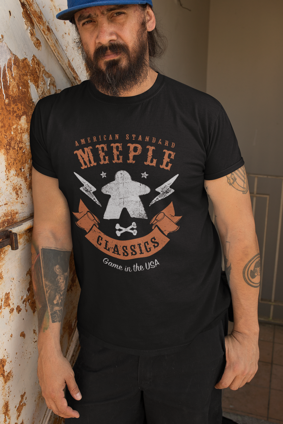 Man wearing a black t-shirt with American Standard Meeple Classics t-shirt, leaning against a wall.
