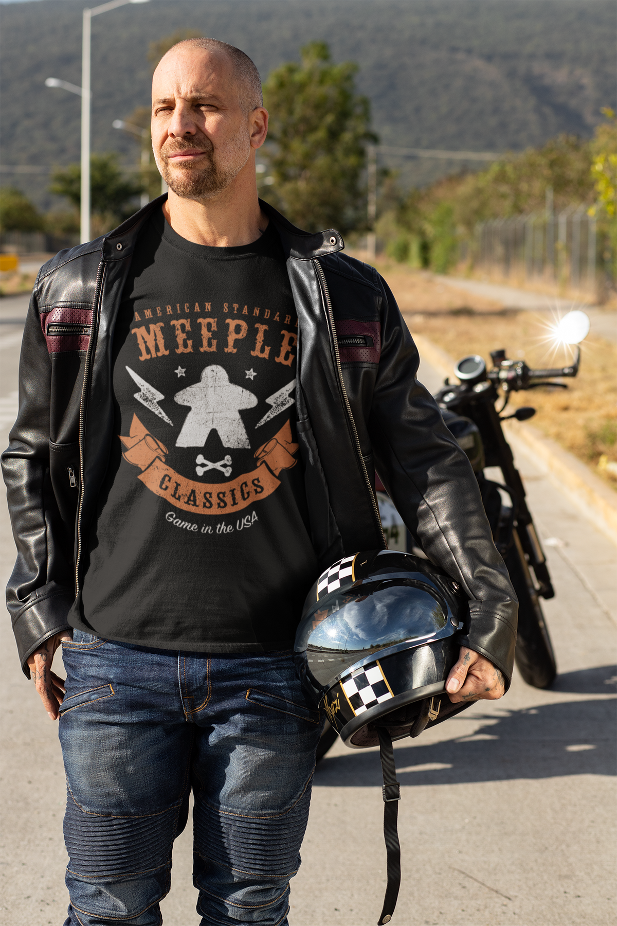 Man in leather jacket holding a helmet on a road with a motorcycle in the background wearing American Standard Meeple Classics T-Shirt