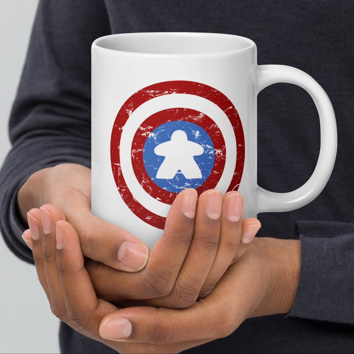Person holding a white mug with our captain meeple design on it, a parody of Captain Americas shield