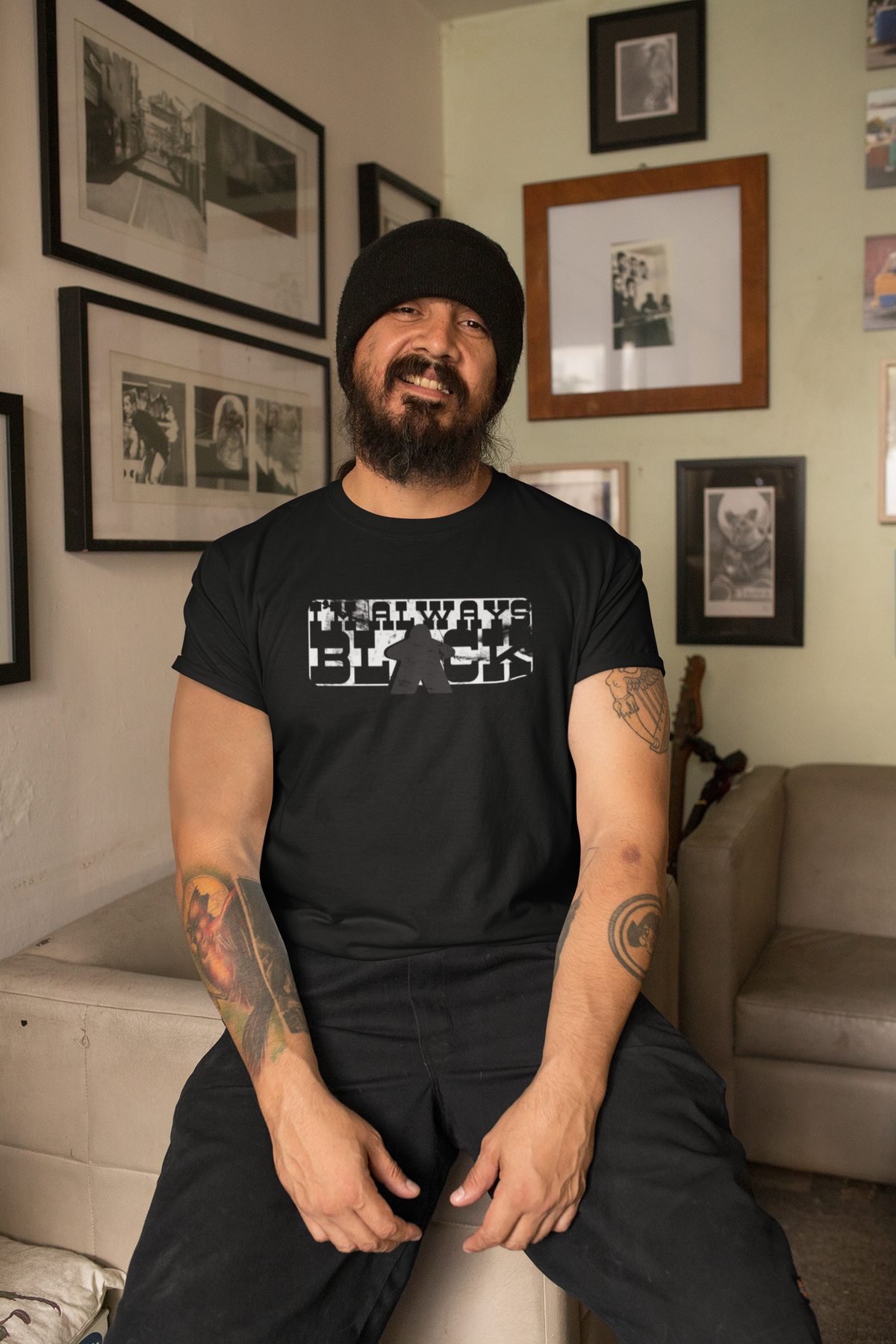 Man wearing a black t-shirt with our I'm Always Black design, sitting in a room with framed pictures on the wall.