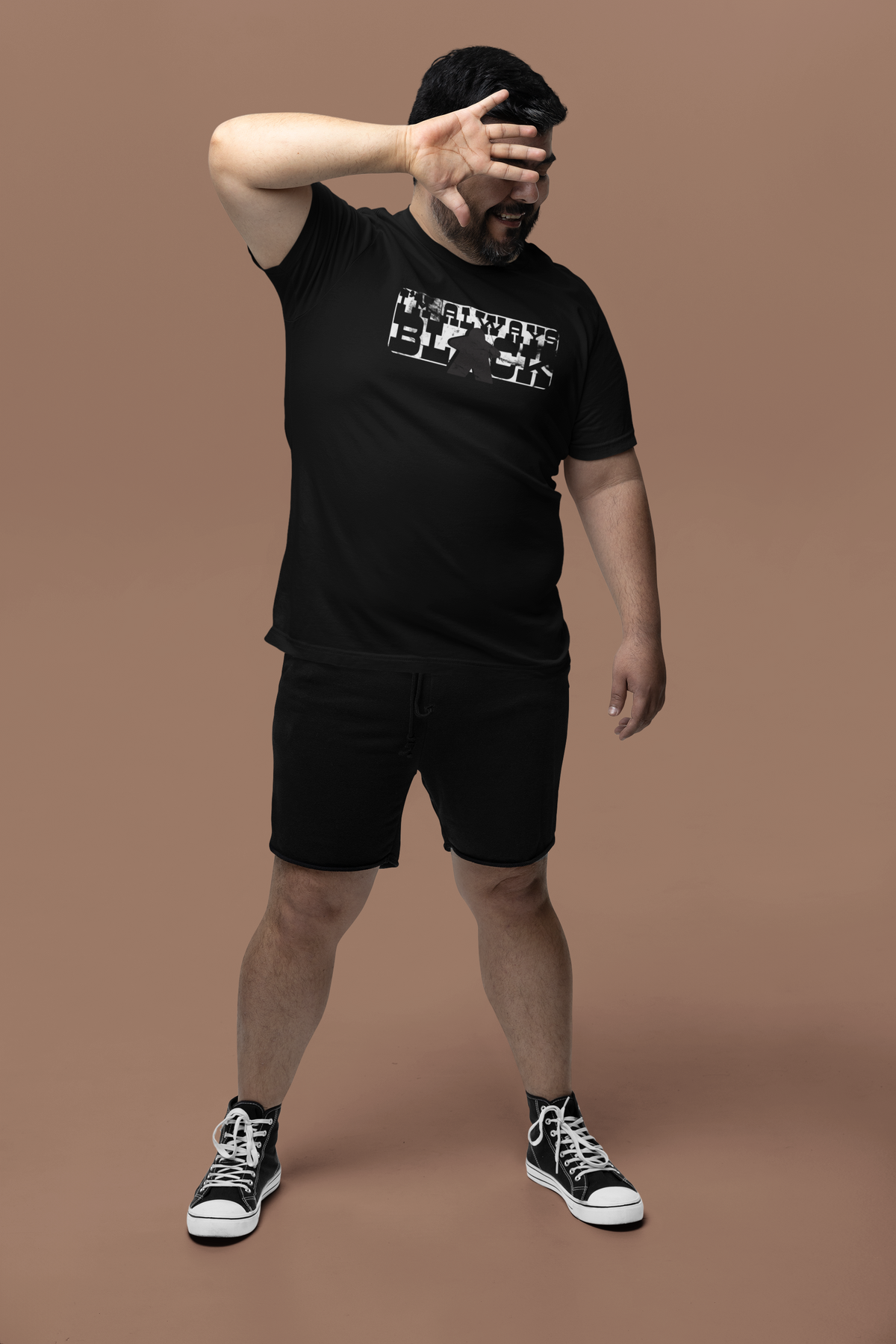 Man wearing a black t-shirt and shorts with our I'm always black design, standing against a brown background.