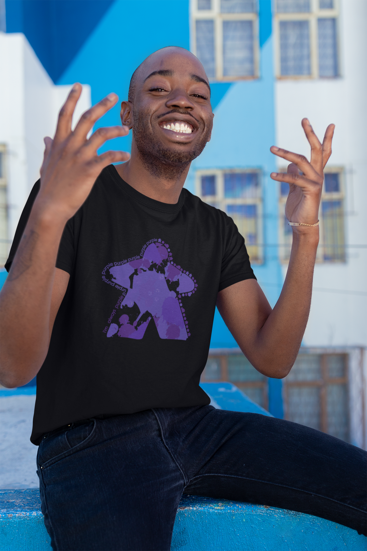Man wearing a black t-shirt with our I'm Always Purple design, sitting against a blue and white building.