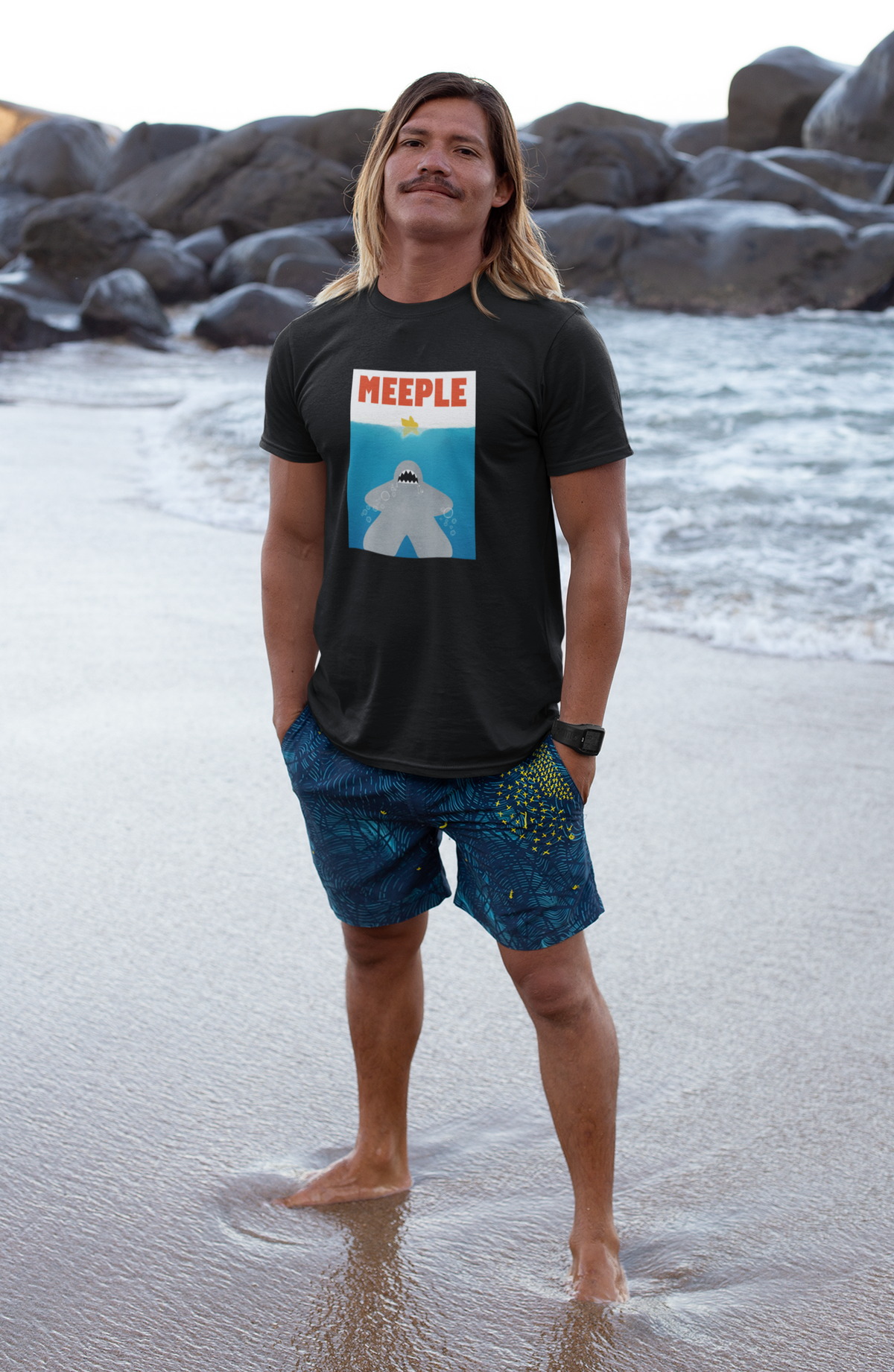 Man standing on the beach with rocks behind him wearing our Meeple Shark Attack t-shirt