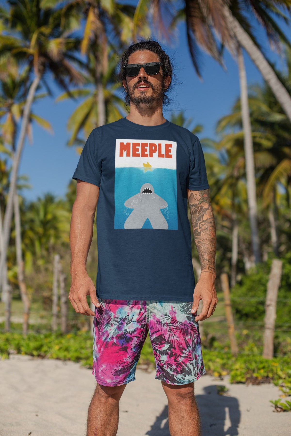 Man standing on the beach with palm trees behind him wearing our Meeple Shark Attack t-shirt