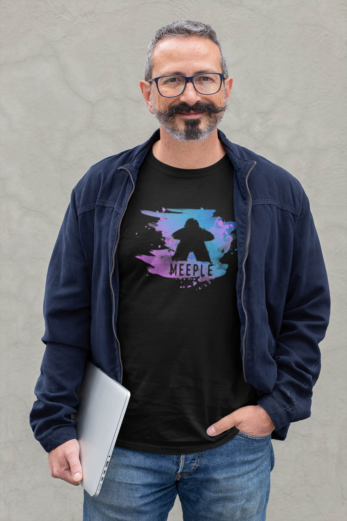 Man standing holding a laptop and wearing a jacket and black t-shirt with our meeple splash design on it.