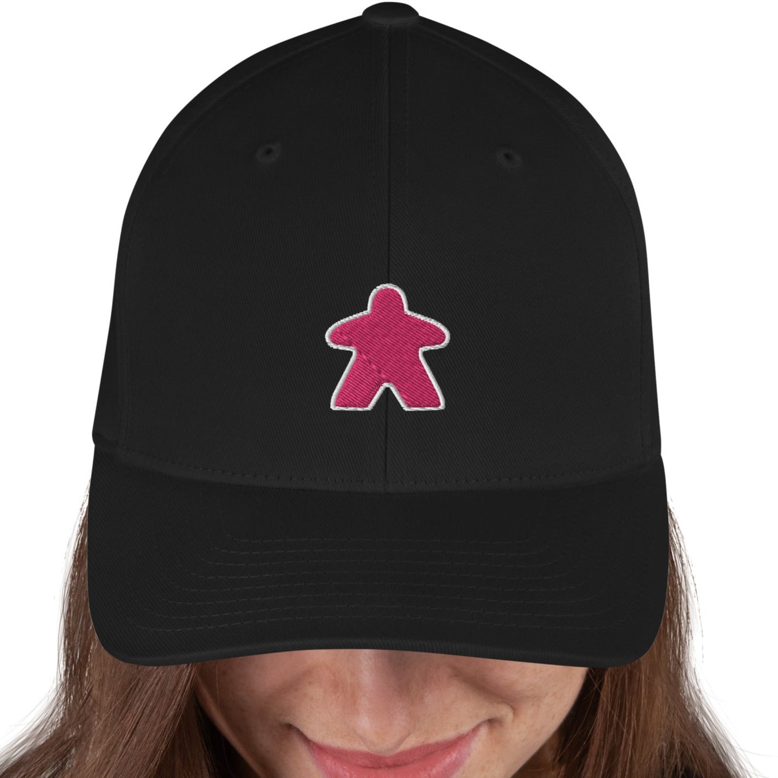 A person wearing a black curved bill hat with a pink embroidered meeple on the front.